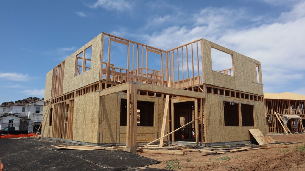 A two-story wooden house frame is under construction on a clear, sunny day.