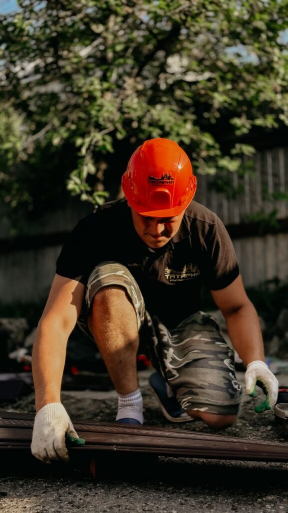 Construction worker in an orange helmet assembles an outdoor structure under bright sunlight.